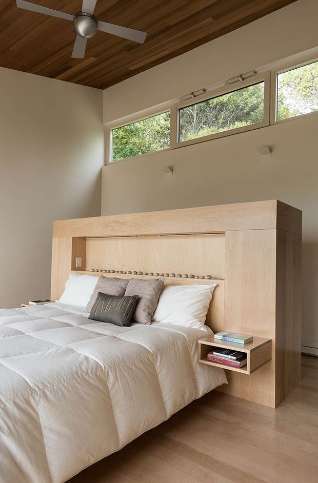 A bright, compact bedroom featuring a pale timber built-in bedhead and surrounding joinery with integrated bedside shelving. The bed is dressed in white bedding with neutral cushions, while high horizontal windows bring in views of greenery. A warm timber-lined ceiling and ceiling fan add natural texture and visual interest.