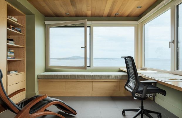 A compact, light-filled home office overlooking the water, with a built-in window seat and storage, a wall-mounted or integrated desk, ergonomic office chair, and open shelving. The room features sage green walls and cabinetry, pale timber ceiling and joinery, large corner glazing, and a calm coastal outlook.