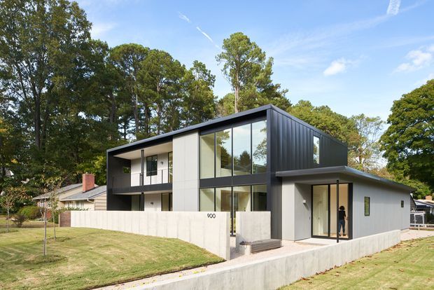 A contemporary two-storey residential house set within a leafy suburban site. The facade combines crisp white wall planes with dark charcoal metal cladding, large floor-to-ceiling glazed sections, recessed balconies, and a low-pitched roof. A concrete boundary wall and paved walkway frame the front garden, while mature trees surround the property.