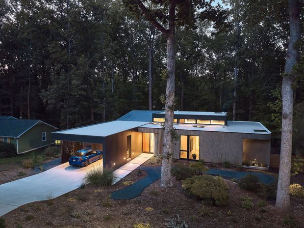 A modern single-storey residence set within a dense wooded landscape, photographed at dusk. The home has a low-profile flat roof with a projecting carport, dark vertical timber-like cladding, extensive warm-lit glazing, and a landscaped front approach. A separate small gable-roofed building is visible to the left, while mature trees dominate the site.