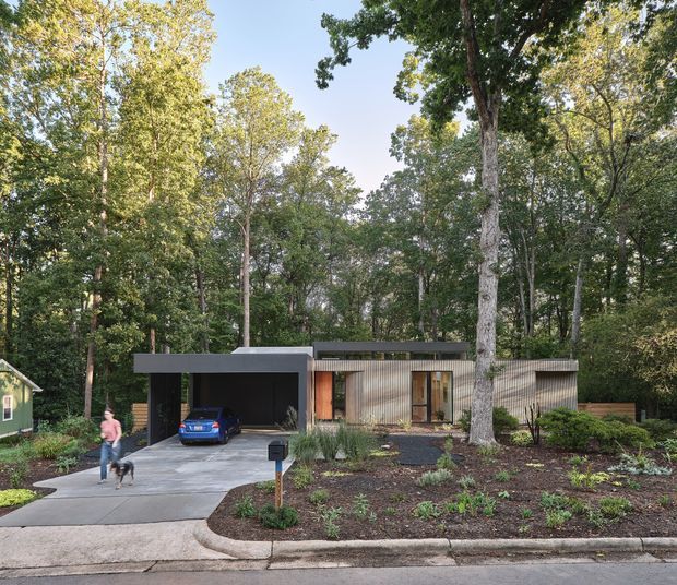 Street-level exterior view of a compact contemporary single-storey home set within a mature wooded landscape. The house has a flat roof, a dark projecting carport or garage, vertical timber-toned cladding, large glazed doors, and a landscaped front garden with a concrete driveway.