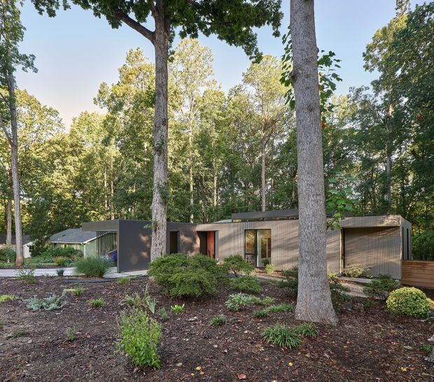 A low, single-storey contemporary residence nestled among mature trees in a landscaped woodland setting. The home has dark horizontal cladding, broad glazed openings, a flat or very low-pitched roofline, and a projecting enclosed carport or garage volume. Layered native-style planting, mulch beds, and a paved approach blend the building into the wooded site.