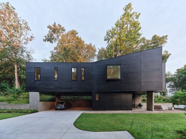 Exterior view of a modern, elevated house with a dark horizontal-clad upper volume, asymmetric pitched roofline, narrow vertical windows, and a larger square window. The upper storey is supported above an open carport and enclosed garage, with a concrete driveway and landscaped lawn in the foreground. Mature trees surround the suburban property.