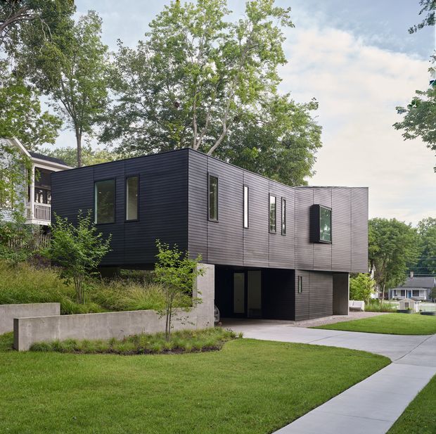 Exterior view of a contemporary two-storey house with strongly cantilevered upper volumes, dark horizontal cladding, narrow vertical windows, and an open ground-level area resembling a carport or sheltered entry. The home sits on a landscaped suburban, slightly sloped site with mature trees, concrete retaining walls, lawn, and a curved concrete walkway.