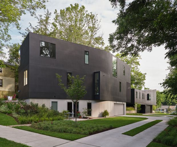 Exterior view of a contemporary multi-storey residence composed of offset geometric volumes. The upper levels are clad in dark vertical ribbed panels with irregularly placed windows, while the lower level is a light neutral finish with garage and entry openings. A landscaped front yard and concrete driveway/walkway surround the house, with mature trees and neighbouring homes visible.
