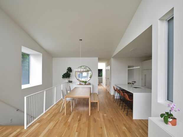 Bright open-plan interior showing a dining area beside a modern white kitchen, with warm timber flooring, a timber dining table, white walls, a linear island with bar seating, pendant lighting, and a stair void with white balustrade. Large windows bring in natural light, and a hallway extends beyond the dining and kitchen areas.