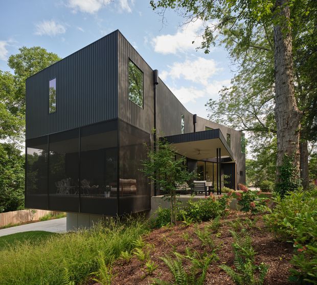Exterior view of a contemporary multi-level house set within a wooded landscaped site. The building has a bold dark vertical-clad facade, tall narrow windows, extensive dark glazing at the lower level, and a projecting covered terrace or entry canopy. Native-style planting, mature trees, and a concrete path surround the home.