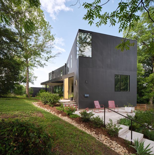 Exterior view of a contemporary multi-level house set among mature trees on a landscaped, sloping site. The building features dark vertically ribbed cladding, large glazed openings, projecting upper-level volumes, and a simple flat or low-pitched roofline. A timber front door opens onto steps and a concrete terrace with two outdoor chairs, while stone edging and native-style planting line the pathway.