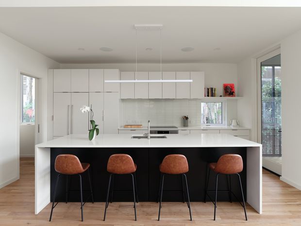 Bright contemporary kitchen with a large central island, four leather-upholstered bar stools, extensive white flat-panel cabinetry, integrated appliances, pale stone benchtops, timber flooring, and dark island cabinetry. The room has abundant natural light from side windows and glazed doors.