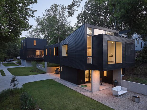 Exterior view of a contemporary multi-level residence composed of offset, box-like volumes clad in dark horizontal siding. Large glazed openings reveal warm illuminated interiors, while concrete supports and a gravel terrace ground the elevated upper volume. A landscaped lawn, concrete paths, mature trees, and a second dark-clad building are visible around the home.