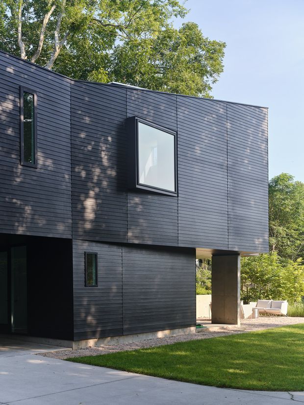 Exterior view of a modern, rectilinear two-storey residence clad in dark horizontal siding. The facade features narrow vertical windows, a projecting square window, a deep recessed ground-floor opening, and a sheltered outdoor terrace or passage supported by a concrete column. The house sits beside a lawn and concrete path, with mature trees surrounding the site.