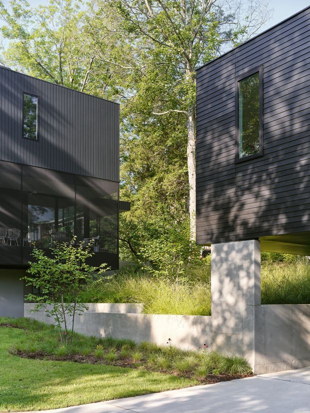 Exterior view of a contemporary elevated residential building composed of two dark-clad volumes separated by a landscaped gap. The structures feature horizontal and vertical dark cladding, large glazed openings, strong cantilevered forms, and exposed concrete supports and retaining walls. The building is set among mature trees and naturalistic grasses with a paved foreground.