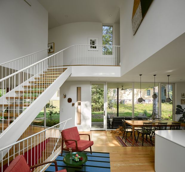Bright double-height residential interior featuring an open stairway with white balustrades, a timber staircase, a mezzanine landing, and a dining and living area arranged around extensive garden-facing glazing. The room includes timber flooring, a red lounge chair, a blue rug, a dining table with pendant lights, and abundant natural light.