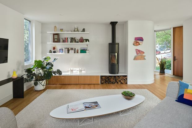 Bright contemporary living room with a freestanding black wood-burning stove set into a white tiled feature wall, floating timber storage cabinetry, open display shelves, and a large oval white coffee table. A grey sofa, indoor plants, artwork, and warm timber flooring create a clean, inviting interior with abundant natural light.