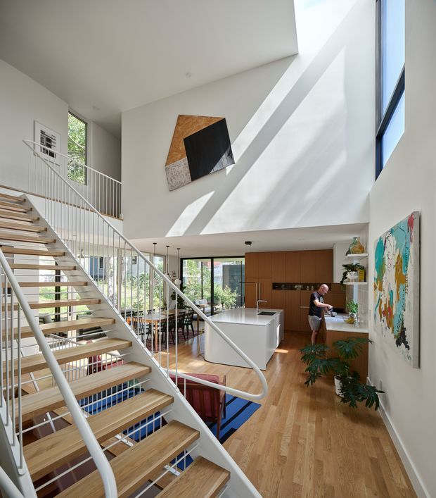 A bright, double-height contemporary interior centered on an open-riser timber staircase with slim white metal railings. The space combines a timber floor, white walls, extensive glazing, a sculptural skylit stairwell, and an open-plan kitchen and dining area. A white island faces warm wood cabinetry, while colourful abstract artwork and indoor plants add visual accents. Strong daylight creates diagonal shadows across the walls.