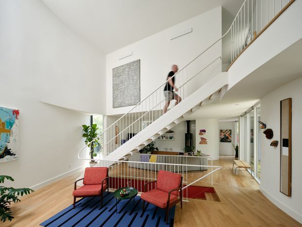 Bright, spacious contemporary open-plan living area with a double-height void and prominent white staircase with slender metal balustrades. The room features pale timber flooring, bold red mid-century-style lounge chairs, a blue patterned rug, indoor plants, artwork, and an upper-level landing visible above. A glazed opening and hallway extend to the right.