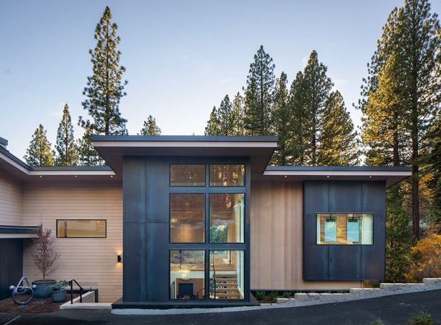 Exterior view of a contemporary single-storey residence set among tall trees. The facade combines horizontal light-coloured cladding with dark metal-clad volumes, broad overhanging flat rooflines, and a tall central glazed entry or stair volume. A paved driveway and landscaped edging are visible in the foreground.