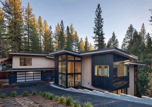 A contemporary single-storey residence set among tall pine trees, featuring a strongly articulated asymmetrical pitched roof, dark vertical cladding, pale horizontal cladding, and extensive floor-to-ceiling glazing around a central entry or living volume. The home includes an attached garage with a glazed sectional door, a dark paved driveway, and landscaped gravel planting beds with ornamental grasses.