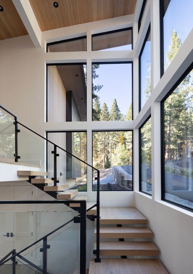 Modern multi-level stairway rising through a bright double-height glazed space. The staircase has open light timber treads, black metal stringers and railings, and clear glass balustrades. Floor-to-ceiling windows frame a wooded outdoor setting, while a timber-lined ceiling and white structural walls create a warm, contemporary interior.
