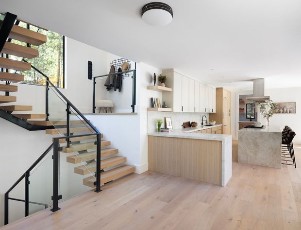 Bright open-plan contemporary home interior featuring a kitchen with a pale stone island and light timber cabinetry, an open timber staircase with black metal and glass balustrades, built-in shelving, white walls, and light timber flooring. The kitchen and dining area extend into the background, with large glazing bringing in natural light.