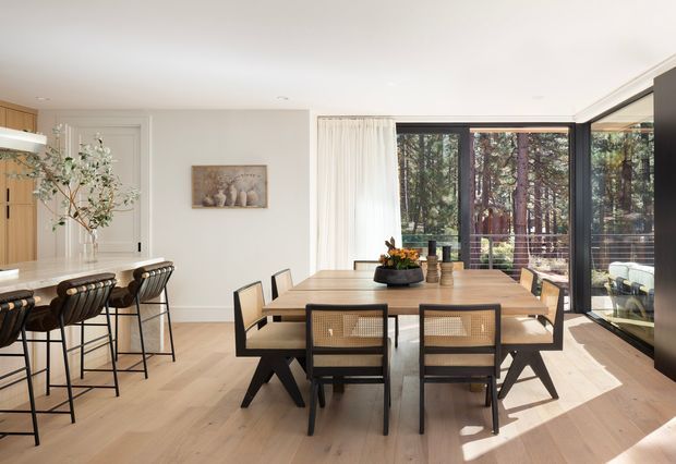 Bright contemporary open-plan dining and kitchen interior with a long light-wood dining table, black-framed woven dining chairs, a kitchen island with bar seating, pale timber flooring, warm wood cabinetry, white walls, and expansive black-framed glazing overlooking trees. The room has abundant natural light and a clean, refined indoor-outdoor connection.