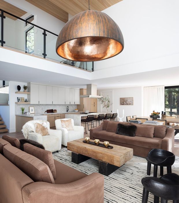 Bright open-plan living and kitchen interior with a double-height ceiling and mezzanine-level railing. The room features warm timber ceiling lining, white walls and cabinetry, a large marble-look island, contemporary bar stools, several brown upholstered sofas and chairs, a substantial rustic timber coffee table, a striped rug, and a dramatic oversized copper dome pendant light.