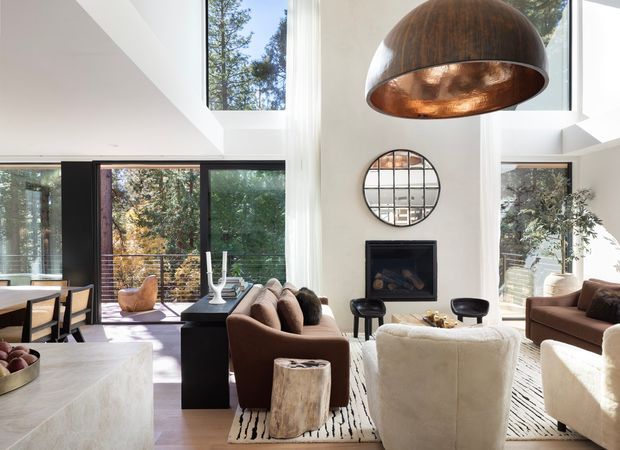Bright, double-height contemporary living room with extensive floor-to-ceiling glazing, a central fireplace, and an oversized copper-toned dome pendant. The space features sculptural upholstered seating, a dark rug with organic stripes, timber flooring, a pale stone kitchen island or counter in the foreground, and views into a leafy setting.