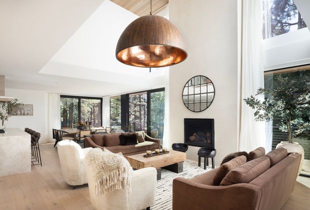 Bright, open-plan living room with floor-to-ceiling glazing, a central fireplace, multiple sofas and armchairs, a timber coffee table, and a large copper pendant light. The space connects visually to a dining area and kitchen, with warm timber flooring and a timber-lined ceiling detail. Large windows overlook trees, while a round grid mirror and soft neutral furnishings add visual interest.