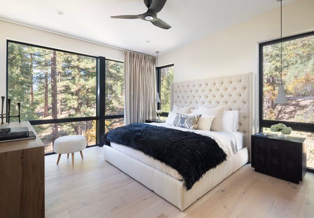 Bright, spacious bedroom with a large upholstered button-tufted bed and tall cream headboard, layered white bedding, and a dark faux-fur throw. The room has pale timber flooring, cream walls, black-framed floor-to-ceiling windows overlooking trees, long neutral curtains, a dark bedside chest, a timber dresser, and a ceiling fan. The styling is luxurious yet relaxed, with abundant natural light and a strong connection to the surrounding woodland.