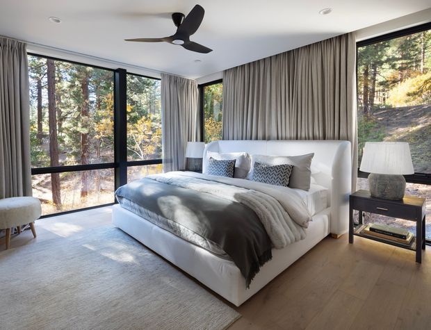 A bright, contemporary bedroom with a large upholstered white bed, layered neutral bedding, patterned accent cushions, and full-height taupe curtains forming a textured feature wall. Expansive black-framed windows overlook a wooded setting, while light timber flooring, a pale area rug, bedside tables, lamps, and a sculptural black ceiling fan complete the calm, refined interior.