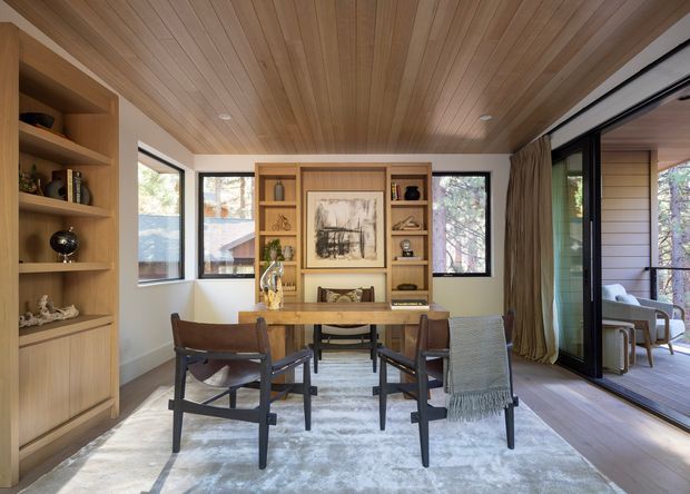 Warm, timber-lined dining and living area with a rectangular dining table, mid-century style armchairs, built-in timber shelving and cabinetry, a central artwork display, and large black-framed sliding glass doors opening to an exterior deck. The space has a timber ceiling, pale walls, wood flooring, and a soft neutral rug, creating a refined, inviting interior with strong indoor-outdoor connection.