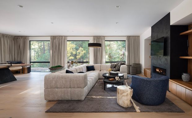 Contemporary open-plan living room with a large cream modular sectional sofa, dark blue lounge chair, layered rugs, round coffee tables, and a sculptural wood side table. Full-height curtained glazing overlooks trees, while a dark stone or plaster fireplace wall incorporates a television and warm timber shelving and cabinetry. The room has a calm, refined, natural palette with light timber flooring and abundant daylight.