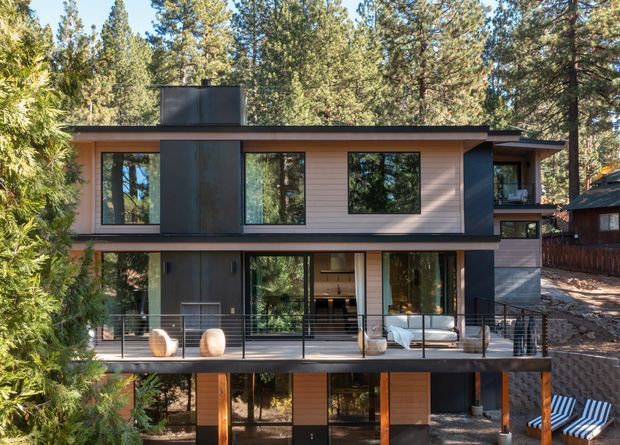 A modern multi-level house set among tall pine trees, viewed from the exterior. The home features clean horizontal lines, dark metal or charcoal cladding, light horizontal wall cladding, extensive floor-to-ceiling glazing, and a large elevated timber deck with slim dark railings. A covered lower level and sheltered outdoor seating are visible beneath the main deck.