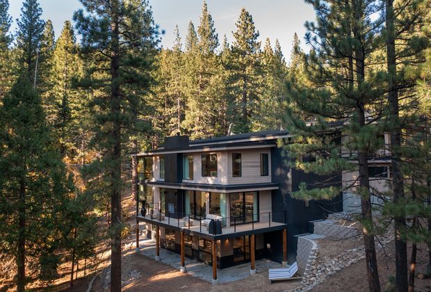 A contemporary multi-level house nestled among dense pine forest, with a dark flat roof, extensive glazing, vertical timber-toned cladding, and broad outdoor balconies or terraces. The elevated structure sits on a sloping, rocky site with retaining walls and stairs, creating a compact woodland retreat.