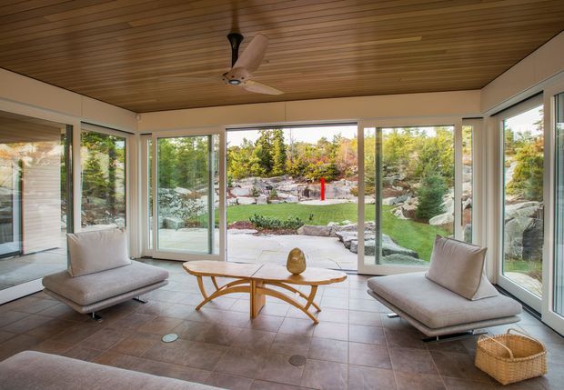Bright glazed sunroom with expansive floor-to-ceiling windows overlooking a landscaped garden and rocky outdoor setting. The room has a warm timber slatted ceiling with a ceiling fan, pale upholstered lounge chairs, a sculptural timber coffee table, and large-format neutral tiled flooring. The open glazing and garden outlook create an airy connection to the outdoors.