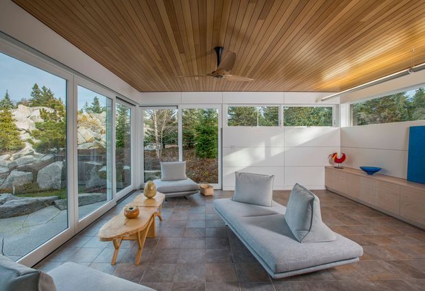 Bright contemporary living room with expansive floor-to-ceiling corner glazing overlooking a landscaped rocky garden. The room has a warm slatted timber ceiling with an integrated ceiling fan, pale built-in cabinetry along the rear wall, large-format dark grey tiled flooring, and minimalist light-grey seating arranged around a low timber coffee table. Linear ceiling lighting and white wall surfaces reinforce the clean, modern aesthetic.