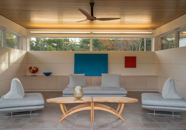 A bright, elongated living room with extensive horizontal glazing, pale built-in cabinetry, and a warm timber slatted ceiling. Two pale upholstered sofas face each other across a sculptural live-edge timber coffee table, with a matching central sofa beneath a large blue artwork and a smaller red artwork. A ceiling fan and linear pendant lighting reinforce the clean mid-century-inspired interior, while trees are visible through the windows.