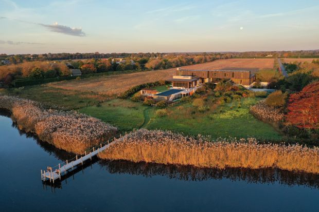 Aerial view of a contemporary low-rise rural residence set among open fields, mature autumn vegetation, and landscaped grounds beside a calm wetland or river. The property includes a long timber-clad building, swimming pool, and a small timber jetty extending into the water.