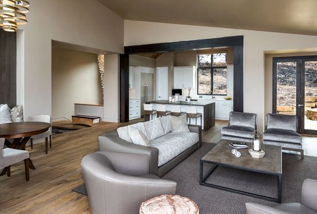 Open-plan contemporary living area with a dining zone, lounge seating and a kitchen visible beyond a wide opening. The room features warm timber flooring, light neutral walls, a dark-framed structural opening around the kitchen, pale upholstered sofas and armchairs, a dark rectangular coffee table, and large windows bringing in natural light. A sculptural pendant hangs above the dining table.