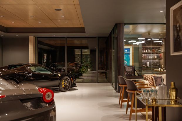 A high-end automotive showroom and lounge with luxury sports cars displayed on a polished light floor. The space features dark timber and glass walls, a warm timber-paneled ceiling with recessed lighting, a bar or dining counter with stools, and a glass-fronted drinks display. The atmosphere is moody, refined, and luxurious.