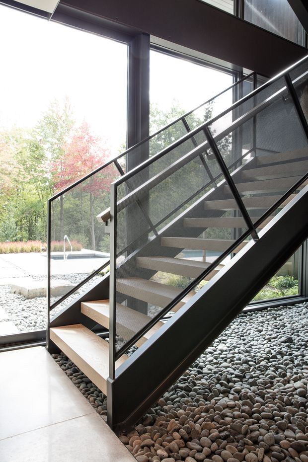 Interior view of a contemporary open-riser staircase with dark metal framing, perforated metal balustrades, and light timber treads. The stair sits within a glazed double-height space, with a pebble-covered floor and landscaped courtyard visible beyond the floor-to-ceiling windows.