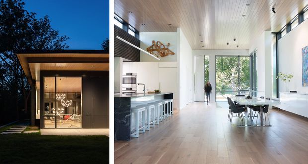A contemporary pavilion-style home shown in a split view: the exterior is a dark, rectilinear volume with a deep flat roof canopy and extensive floor-to-ceiling glazing, while the interior is an open-plan kitchen, dining and living space with a light timber-lined ceiling, pale timber flooring, white walls and abundant natural light. The interior includes a kitchen island with stools, a dining table, large windows and built-in cabinetry.