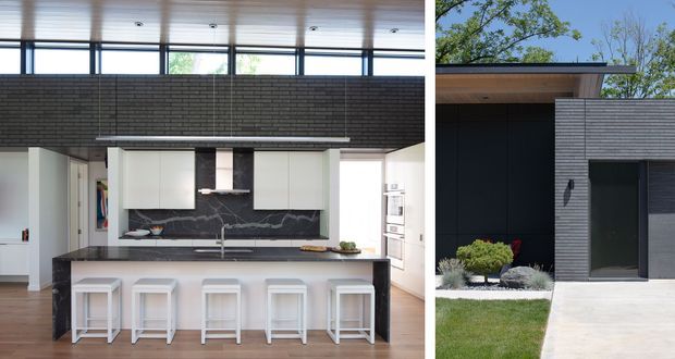 Split image showing a contemporary kitchen interior on the left and a modern single-storey house exterior on the right. The kitchen features a large island with four white stools, white flat-panel cabinetry, a dark veined stone splashback and matching dark island sides, light timber flooring, clerestory glazing, and integrated appliances. The exterior has dark horizontal brick cladding, a broad flat roof, dark window and wall surfaces, and a simple concrete path with landscaped planting.