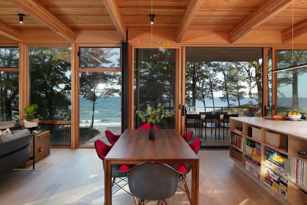Open-plan dining and living area with a timber-beamed ceiling, extensive timber-framed glazing, and direct views toward the ocean and shoreline. A dark wood dining table with red chairs occupies the centre, while built-in shelving and a kitchen or serving counter line the right side. The room has a warm natural-material palette and strong indoor-outdoor connection.