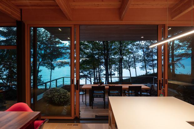 Warmly lit contemporary dining and kitchen interior opening onto an outdoor deck, with extensive dark-framed glazing overlooking trees and a vivid blue waterfront. The room features a timber-lined ceiling with exposed beams, timber-framed sliding doors, a large pale kitchen island, and an outdoor dining setting beyond.