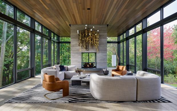 A spacious contemporary living room enclosed by floor-to-ceiling black-framed glazing on three sides, overlooking dense landscaped greenery. The room features a curved cream sectional sofa, sculptural tan lounge chairs, a low coffee table, a monochrome striped rug, a central fireplace set into a grey stone or concrete feature wall, and an abstract sculptural artwork. Warm timber boards line the ceiling, while pale timber flooring and abundant natural light create an airy, refined interior.