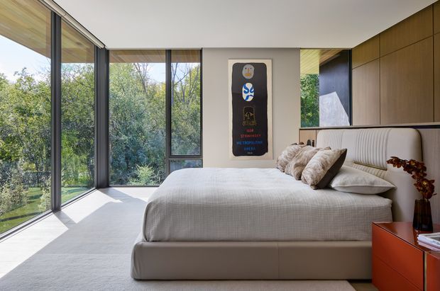 Contemporary bedroom with a low upholstered platform bed, curved channel-tufted headboard, neutral textured bedding, and warm timber built-in cabinetry. Floor-to-ceiling corner glazing opens the room to leafy outdoor views, while a sculptural wall artwork and rust-coloured bedside cabinet add visual interest. The space is bright, refined, and connected to nature.