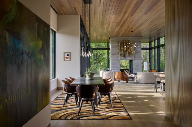 A sophisticated open-plan dining and living area with a timber-lined ceiling, expansive black-framed glazing, and strong indoor–outdoor visual connection to surrounding greenery. The dining zone features a dark timber table, sculptural upholstered dining chairs, and a patterned area rug, while the adjoining lounge includes contemporary sofas and armchairs. Large-scale artwork, decorative pendant lighting, a stone-textured feature wall, and layered natural materials create a warm, refined atmosphere.