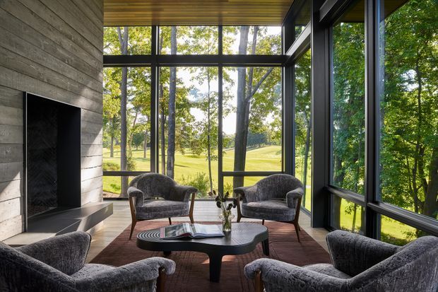 A refined, glass-walled sitting room immersed in a wooded landscape. Four upholstered lounge chairs surround a low sculptural coffee table on a dark rug, with a large built-in fireplace clad in horizontal timber boards along the left wall. Floor-to-ceiling black-framed glazing provides expansive views of mature trees and open lawn. The ceiling features warm timber lining, creating a warm, modern retreat.