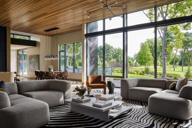 A spacious contemporary living room with a large sculptural sectional seating arrangement, low white rectangular coffee table, and black-and-white patterned rug. The room opens to a dining area and features extensive floor-to-ceiling black-framed glazing overlooking a landscaped green setting. A warm timber-lined ceiling, integrated linear lighting, modern ceiling fan, and sculptural dining pendant add strong architectural character.